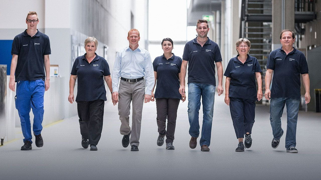 Row of 7 Ensinger employees walking in a factory hall towards the observer